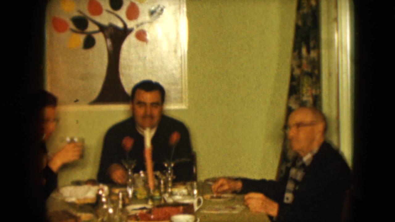 1961 Nevada home-movie frame of three men gathered at a table with cups, glassware, candle, and wall art in a private interior.