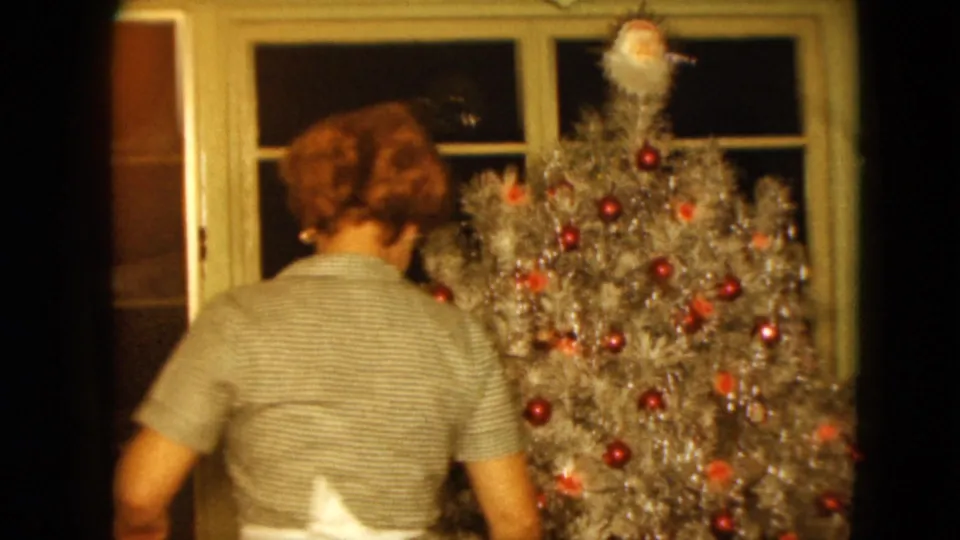 1961 Nevada home-movie frame showing a woman decorating or standing near a silver tinsel Christmas tree with red ornaments.