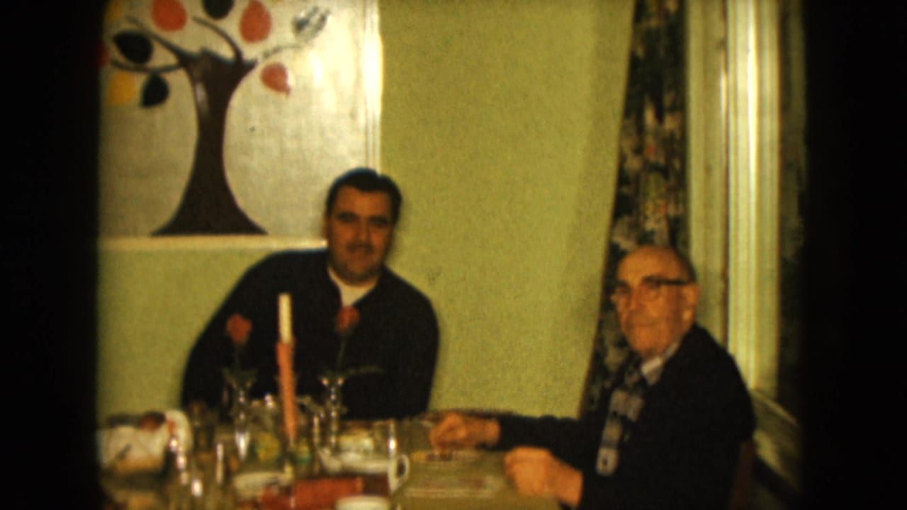 In a 1961 Nevada home interior, a younger man and an older man sit at a table with coffee cups, glassware, flowers, and a wall tree painting.