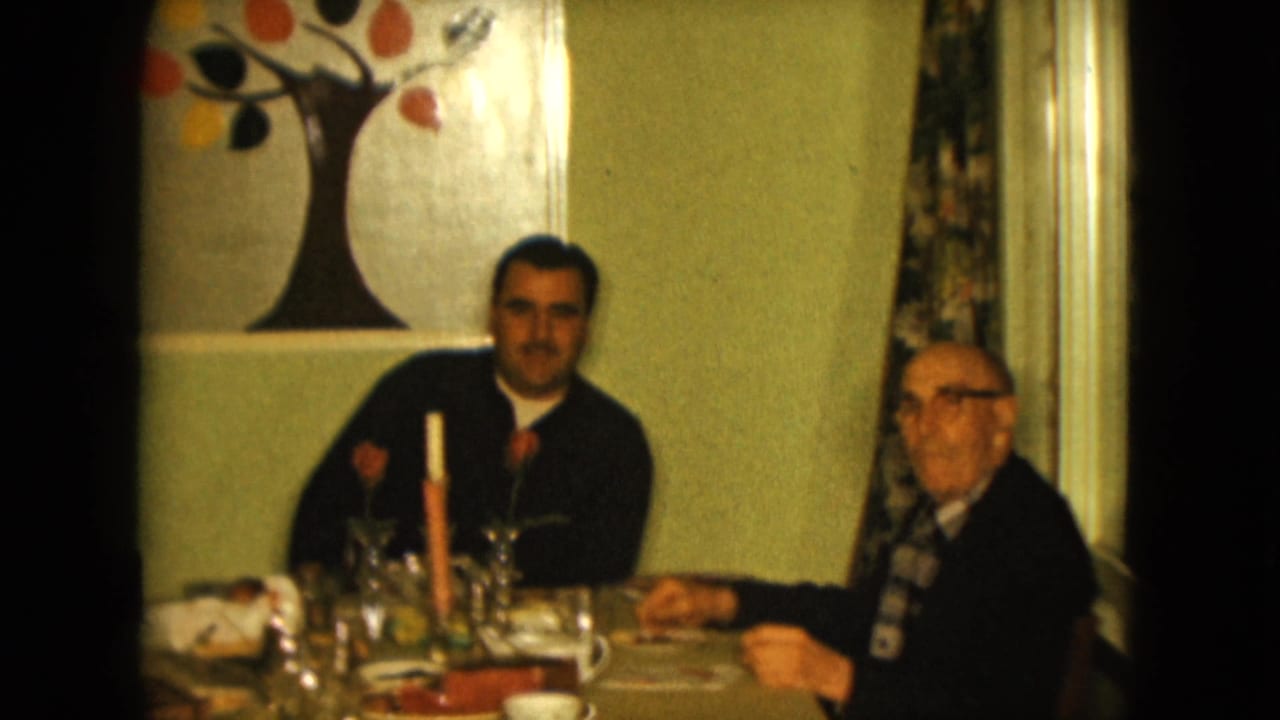 A younger man and older man in a Nevada living room face the camera while seated at a table with cups, glassware, a candle, and wall art.