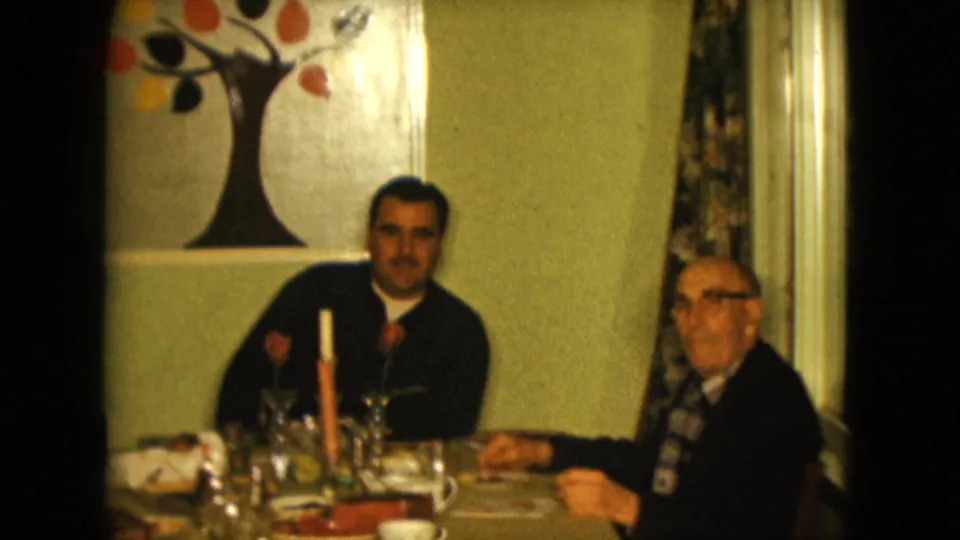 A younger man and older man in a Nevada living room face the camera while seated at a table with cups, glassware, a candle, and wall art.