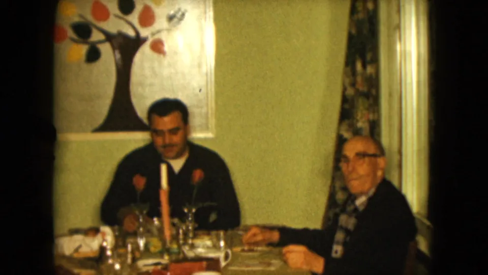 In the 1961 home-movie scene, the younger man looks down at the table as the older man sits beside him with cups and glassware nearby.