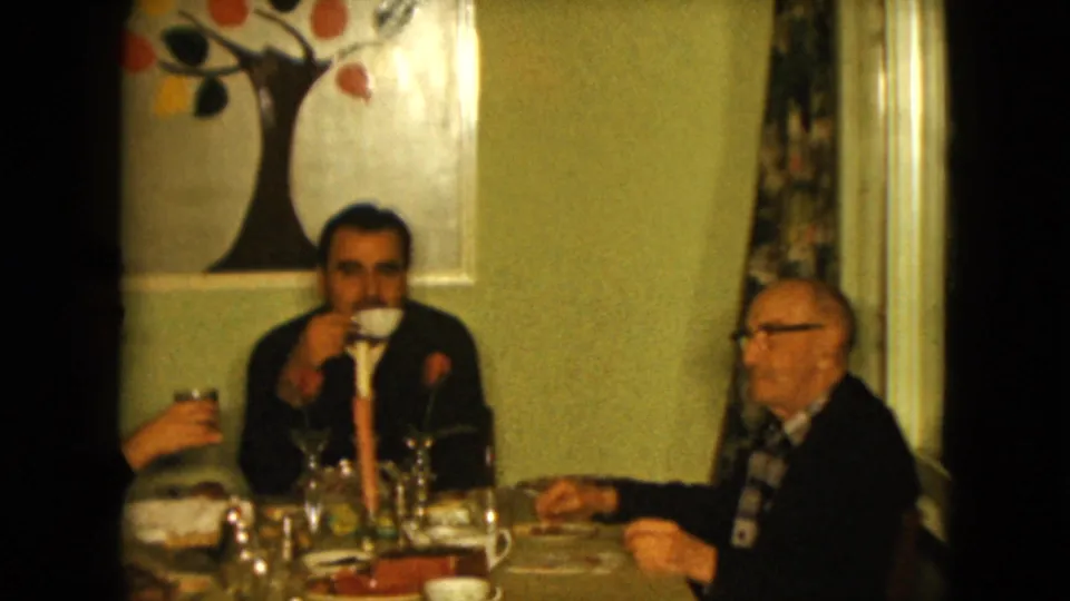 The younger man lifts a cup to drink while the older man remains seated beside him in the Nevada living room setting.