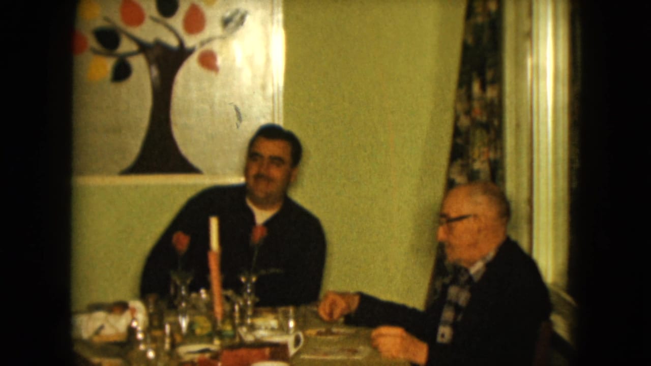 Two men seated at a dining table with glasses, plates, a candle, and wall art behind them.
