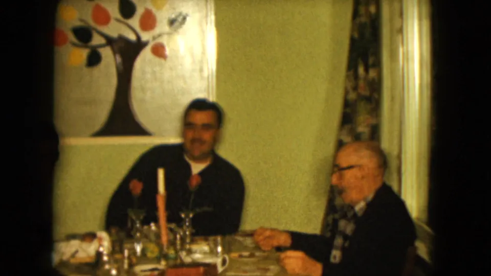 1961 Nevada home-movie frame of two men at a family dinner table, with the older man in profile and the younger man near painted wall decor.