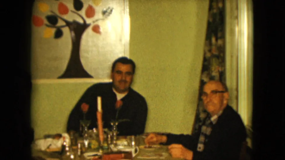 1961 Nevada home-movie frame showing an older man looking toward the camera, with a younger man and a set dining table beside him.