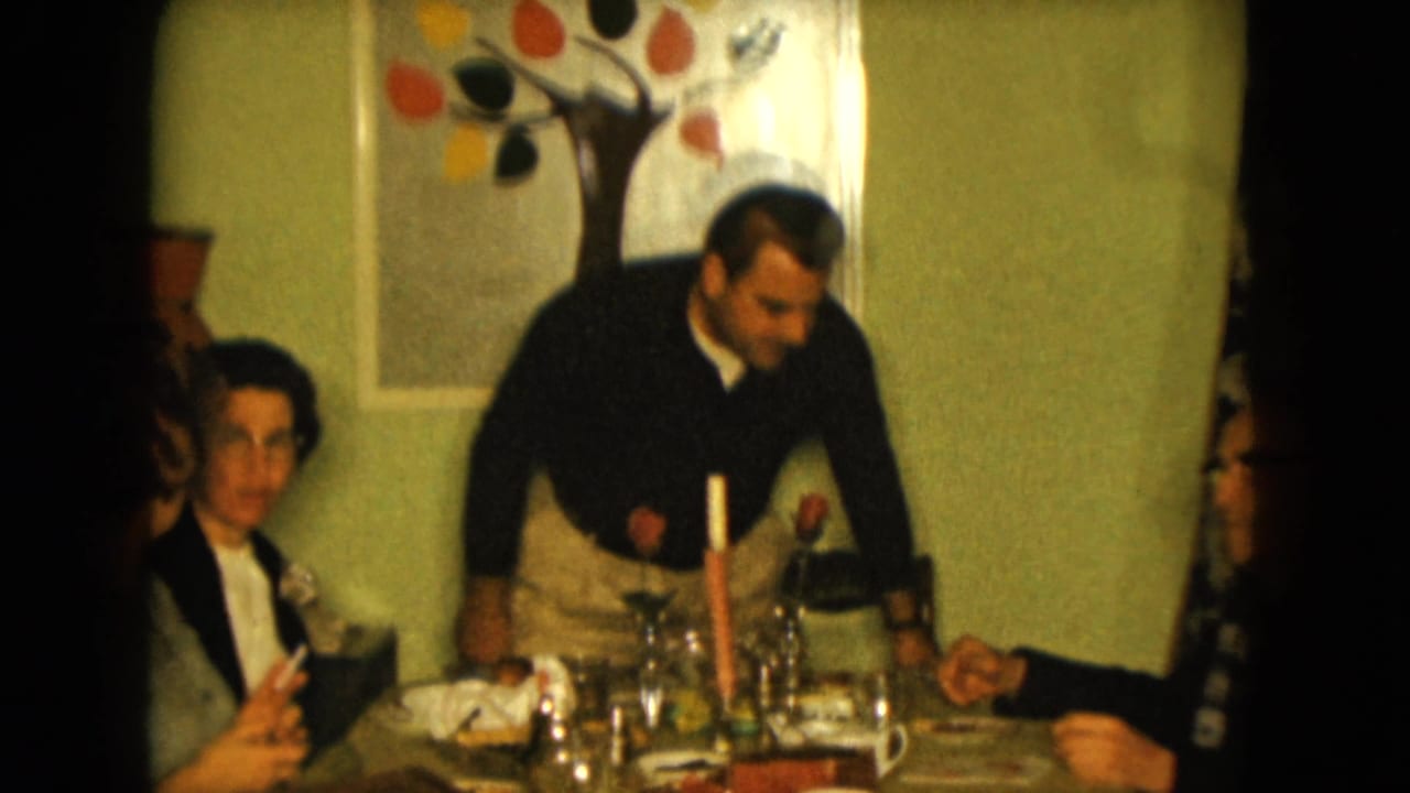 1961 Nevada home-movie frame of adults at a dining table, with a standing man, seated woman, glasses, candle, roses, and tree artwork on the wall.