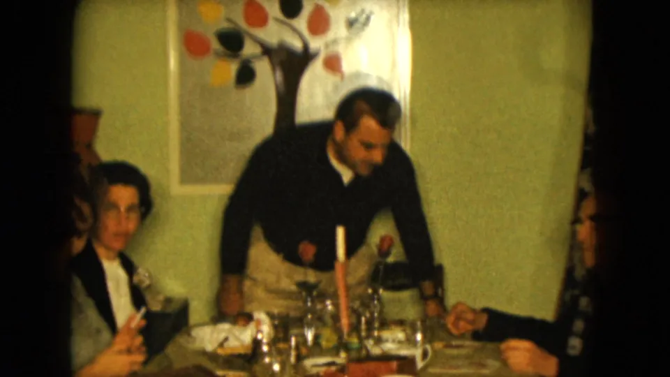 1961 Nevada home-movie frame of adults at a dining table, with a standing man, seated woman, glasses, candle, roses, and tree artwork on the wall.
