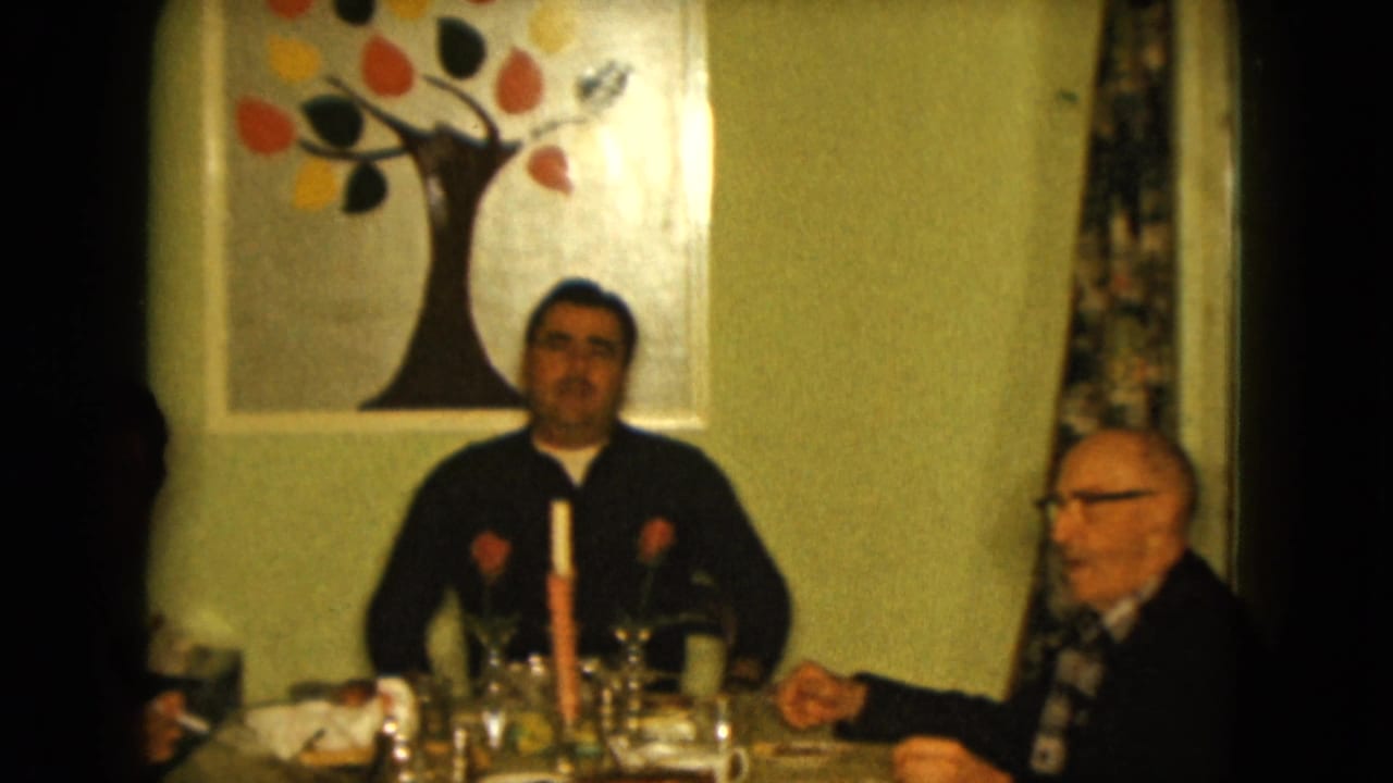 Two men sit at a dining table with glasses, flowers, and a candle.