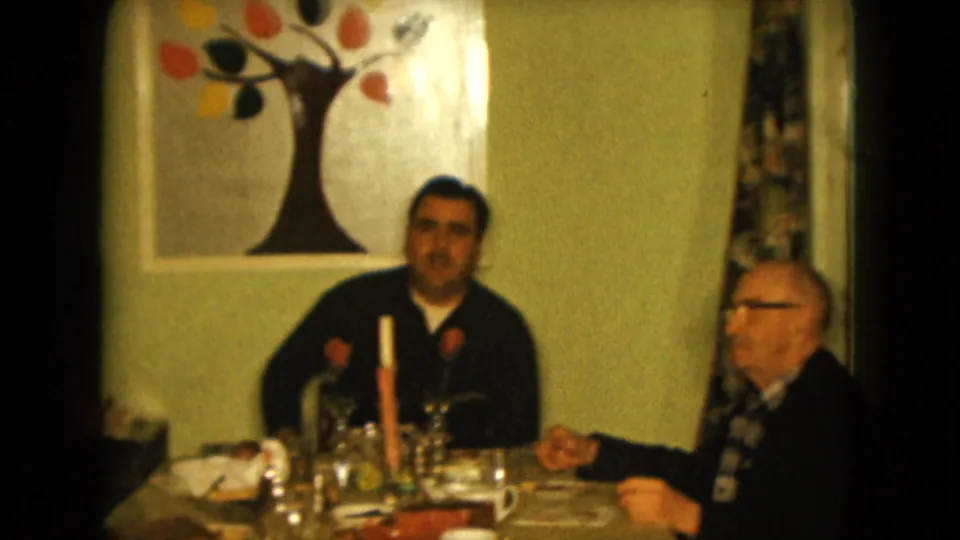 1961 Nevada home interior with seated men at a set table, visible candle, glassware, red roses, patterned curtains, and a tree painting.