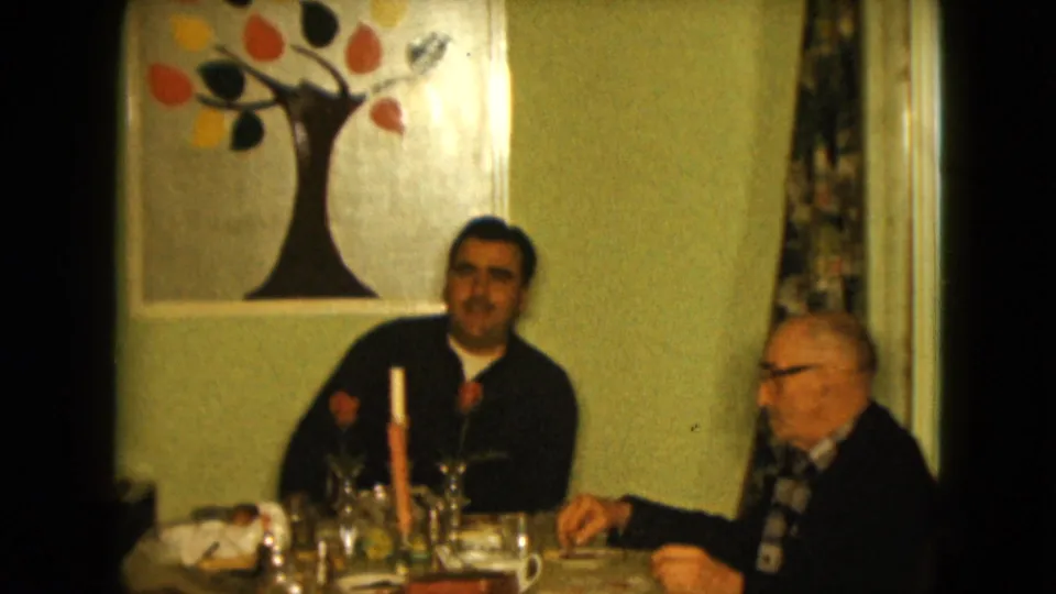 1961 Nevada home-movie frame of two men seated beside a dining table, with a candle, glassware, roses, curtains, and autumn-colored tree artwork.