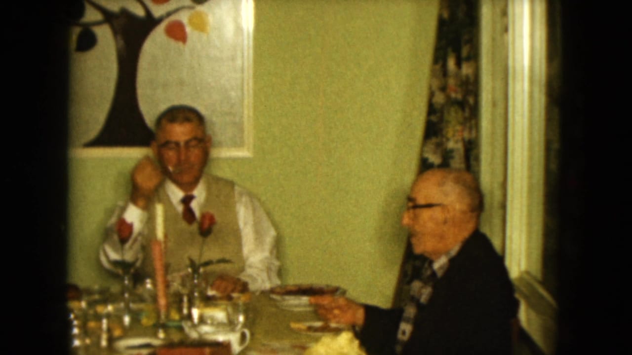 1961 Nevada home-movie frame showing two older men seated at a decorated dining table in a warm private interior.