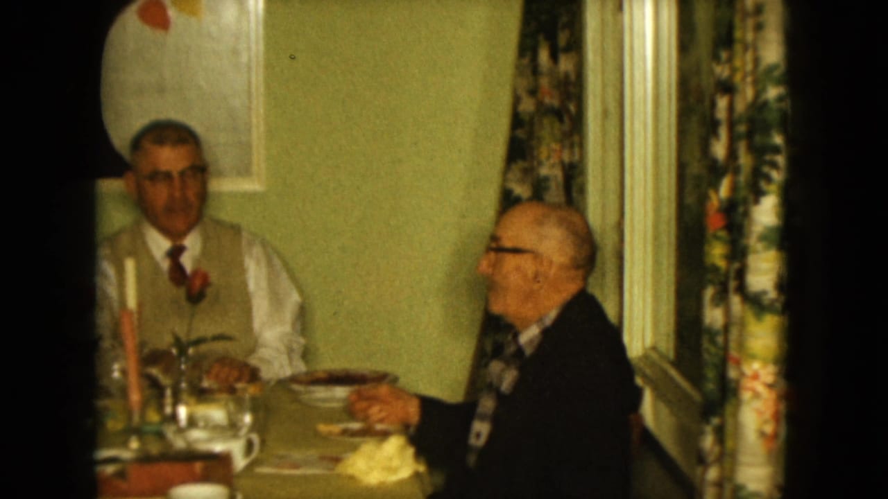Two men sit at the table while one gestures near his face beside candles, glasses, and floral curtains.