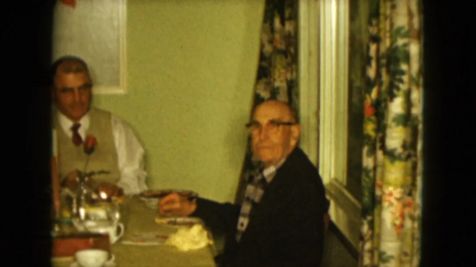 1961 Nevada home-movie frame of two older men at a dining table, with one looking toward the camera in a private room.