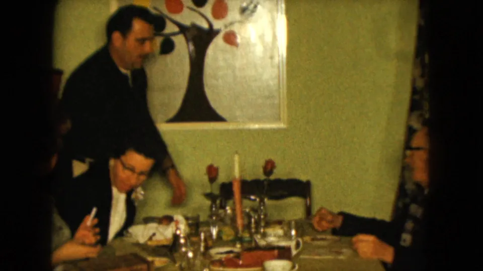 1961 Nevada interior frame showing a standing man behind a seated man at the dining table, with roses and wall art visible.