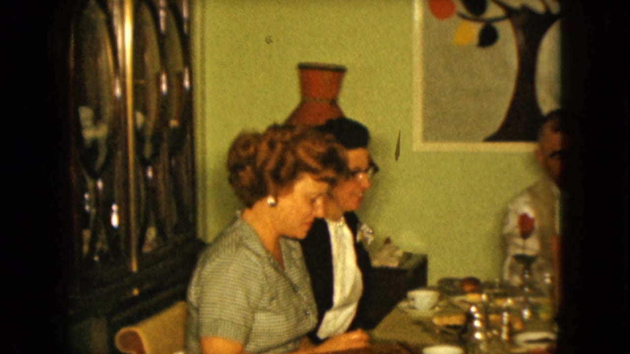 In a 1961 Nevada 8mm home movie frame, adults sit around a dining table beneath tree artwork on a green wall.