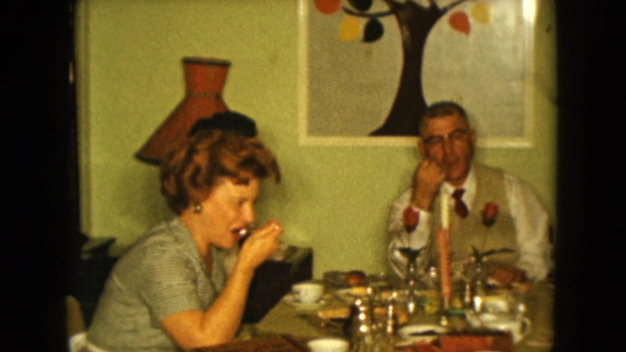 Woman eating with a spoon at a dinner table while a man in a vest sits nearby.