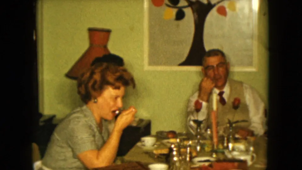 A woman in a checkered dress eats from a spoon as adults sit at a table in the 1961 Nevada home-movie scene.