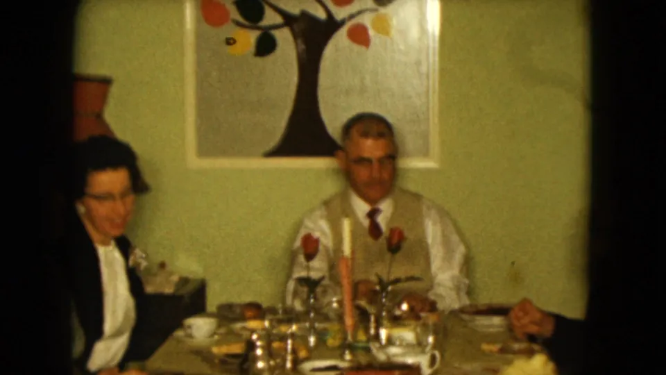 Adults at a 1961 Nevada family dinner sit around a set table with glassware, candles, and domestic wall decor.