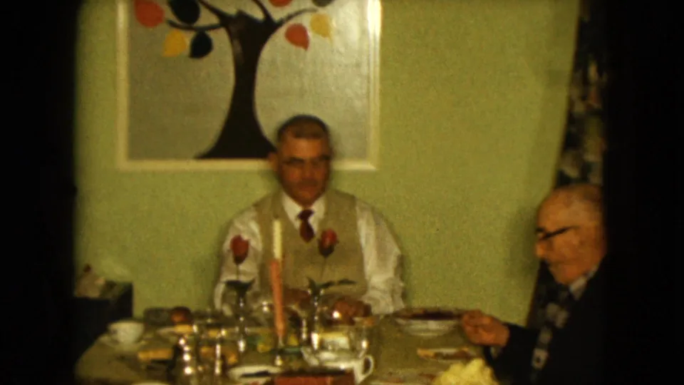 Two men are visible at a family dinner table in a 1961 Nevada home movie, with candles and a tree artwork behind them.