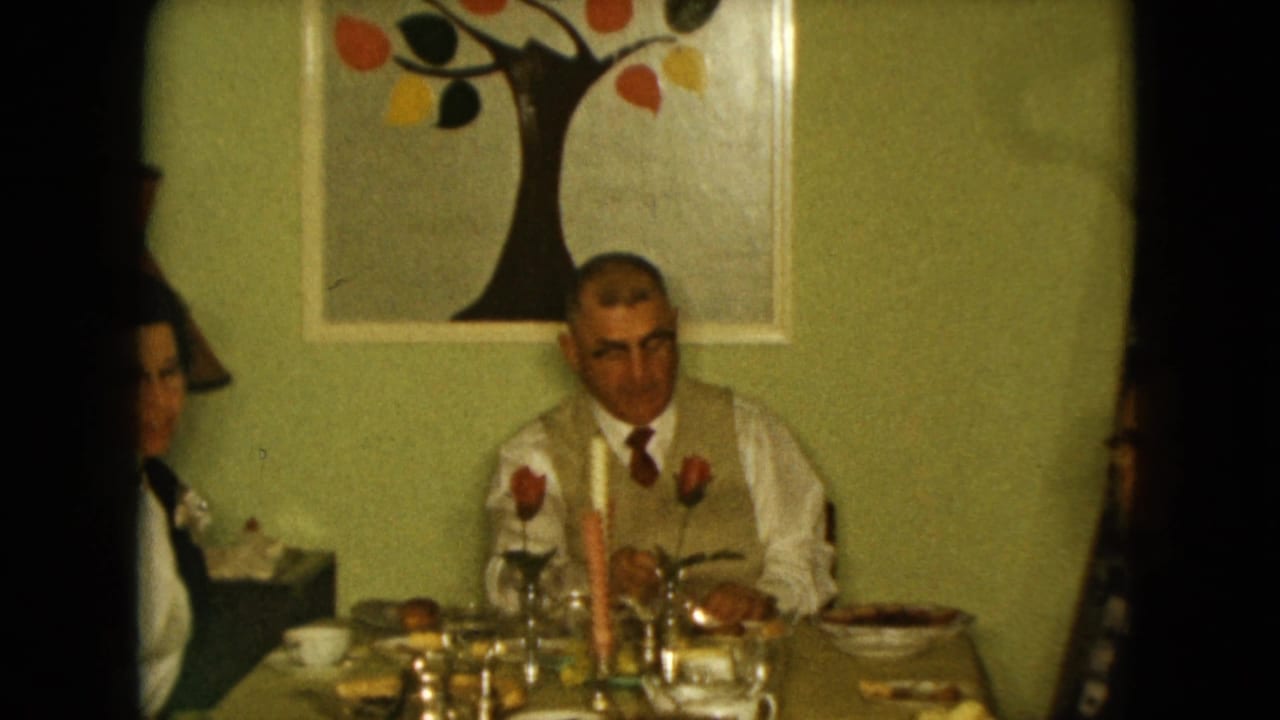 1961 Nevada home-movie frame of adults at a family dinner table, with roses, a tall candle, cups, dishes, and abstract tree wall art.