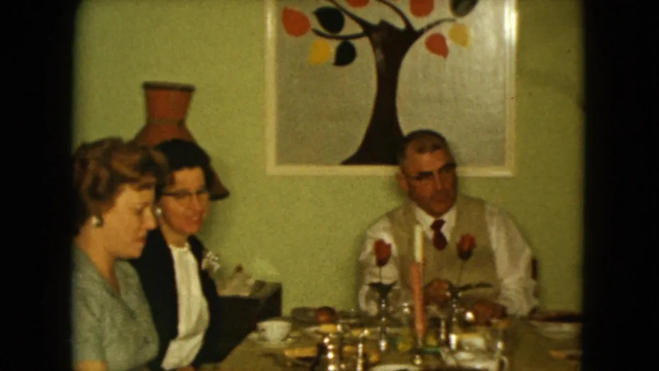 1961 Nevada dinner gathering with two women in profile and a man seated near table settings, roses, candle, and wall art.