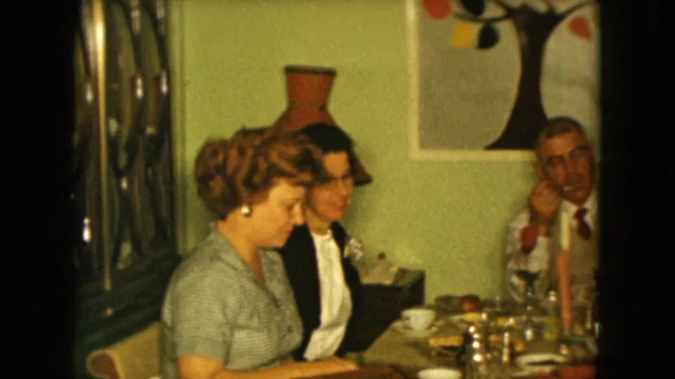 1961 Nevada home interior showing two women at a dining table, a reflective cabinet, tableware, and a man partly visible at right.