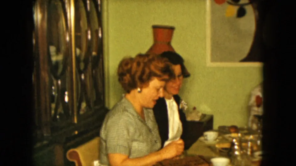 1961 Nevada home-movie frame of two women seated at a family dinner table near a reflective cabinet and mid-century wall decor.