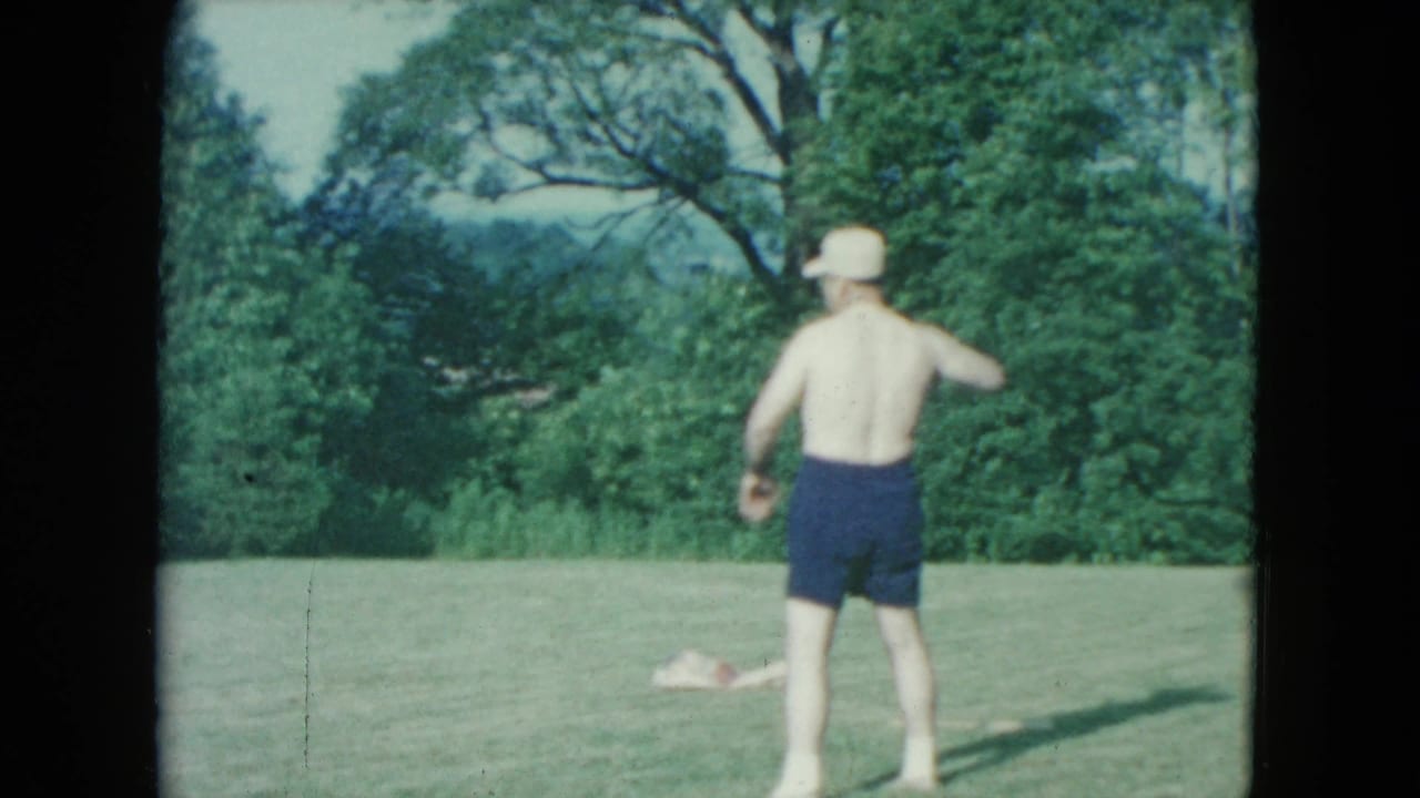 In 1961 Nevada home-movie footage, a shirtless man in a white hat and dark shorts stands outdoors on grass near leafy trees.