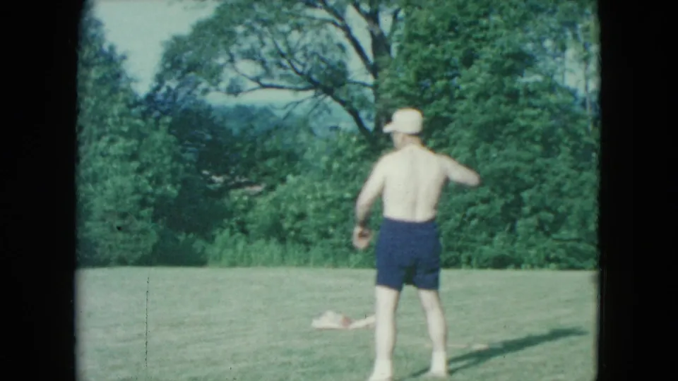 In 1961 Nevada home-movie footage, a shirtless man in a white hat and dark shorts stands outdoors on grass near leafy trees.