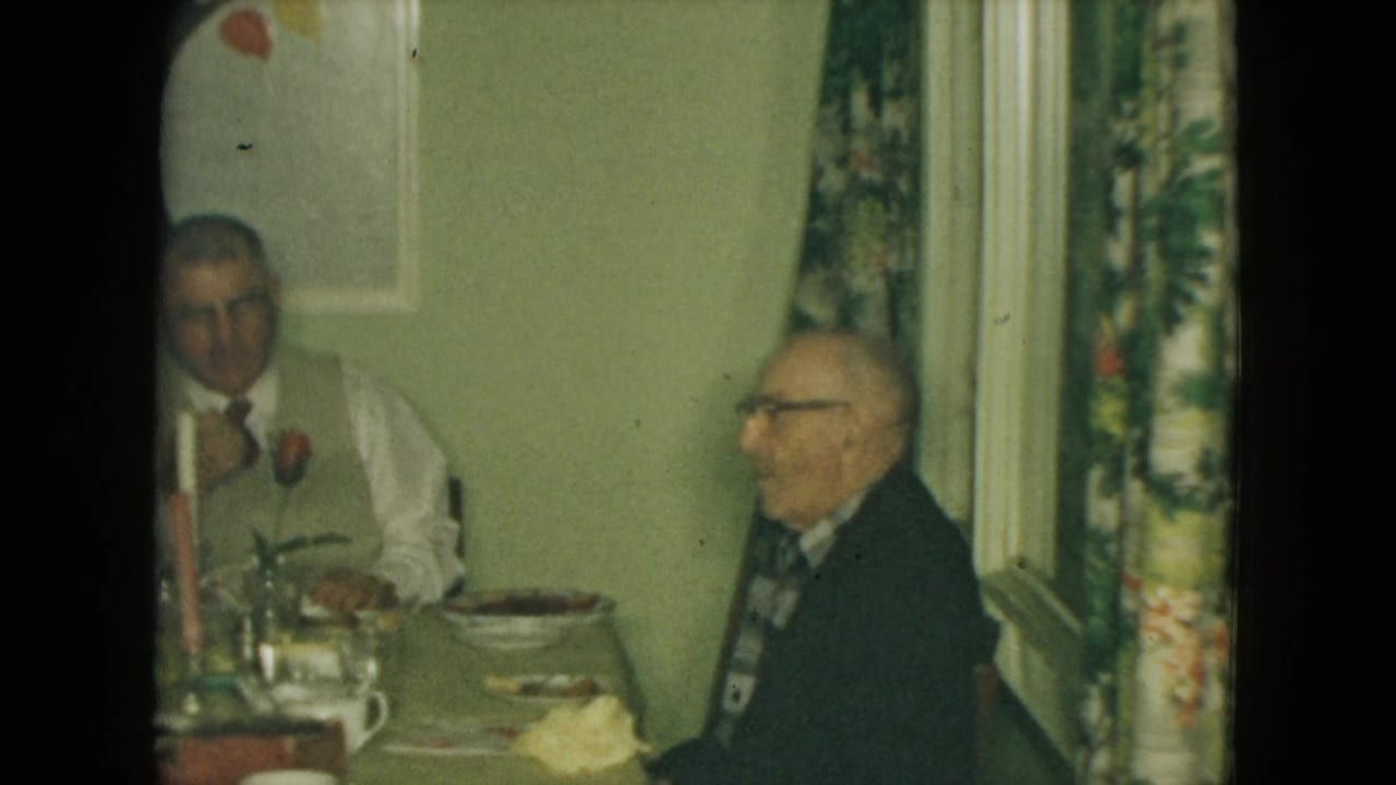 Two elderly men sit at a dining table beside floral curtains and a wall painting.