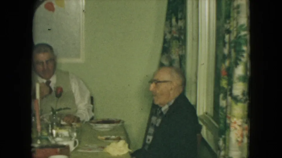 Two elderly men sit at a home dining table in 1961 Nevada footage, with candles, plates, floral curtains, and wall art visible.