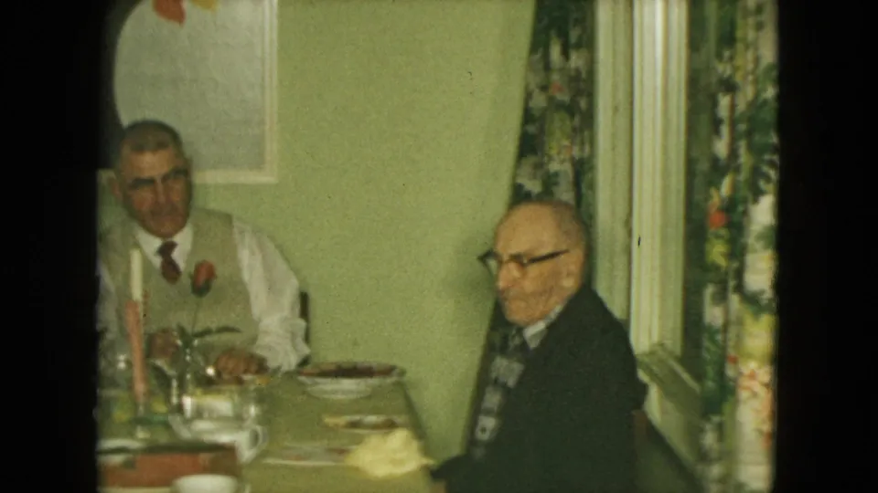 The indoor 1961 Nevada home-movie scene shows two elderly men seated at a dining table with candles, dishes, and curtains nearby.