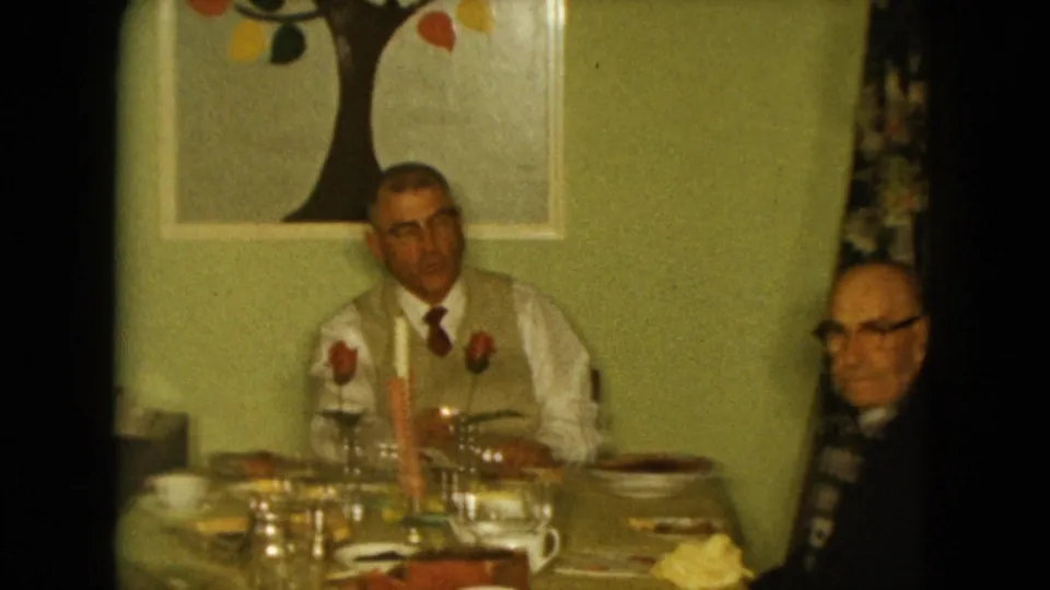 Two elderly men sit at a candlelit dining table in a private Nevada home interior, with wall art and place settings visible.