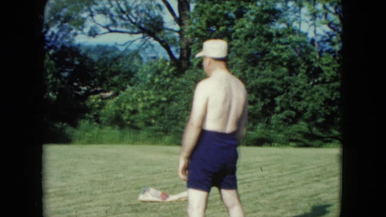 1961 Nevada home-movie frame showing a shirtless man in blue shorts and a white cap facing away on a green lawn.