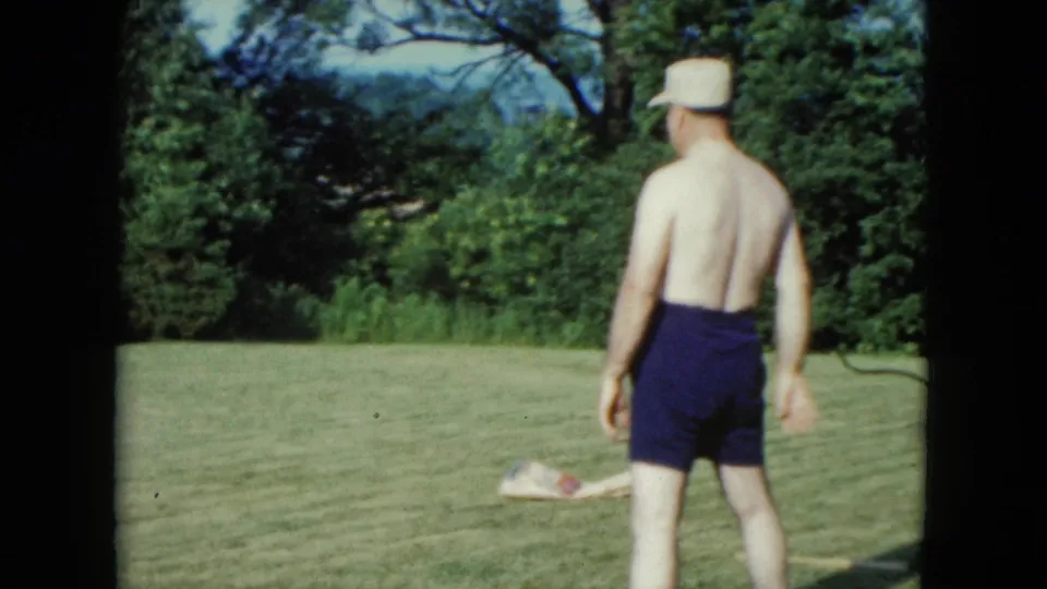 1961 Nevada home-movie frame showing a shirtless adult man in a light hat and dark swim trunks standing on the backyard lawn.