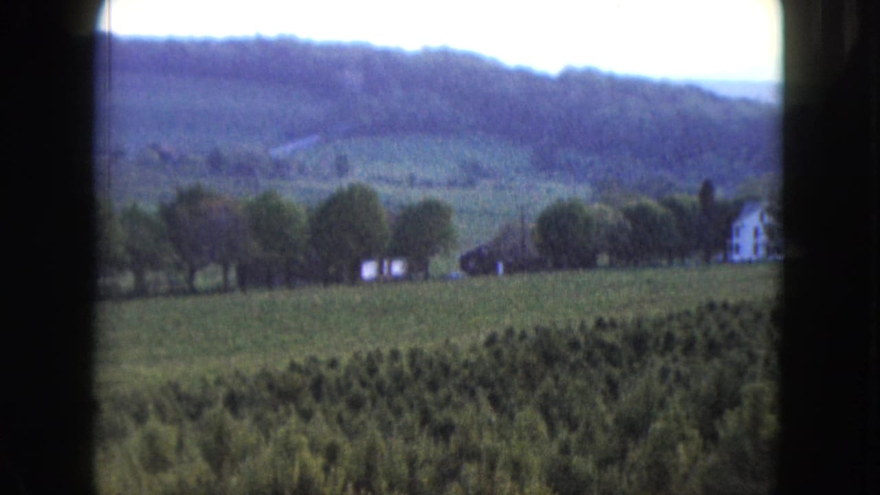 Green fields and trees lead toward a white farmhouse beneath wooded hills.