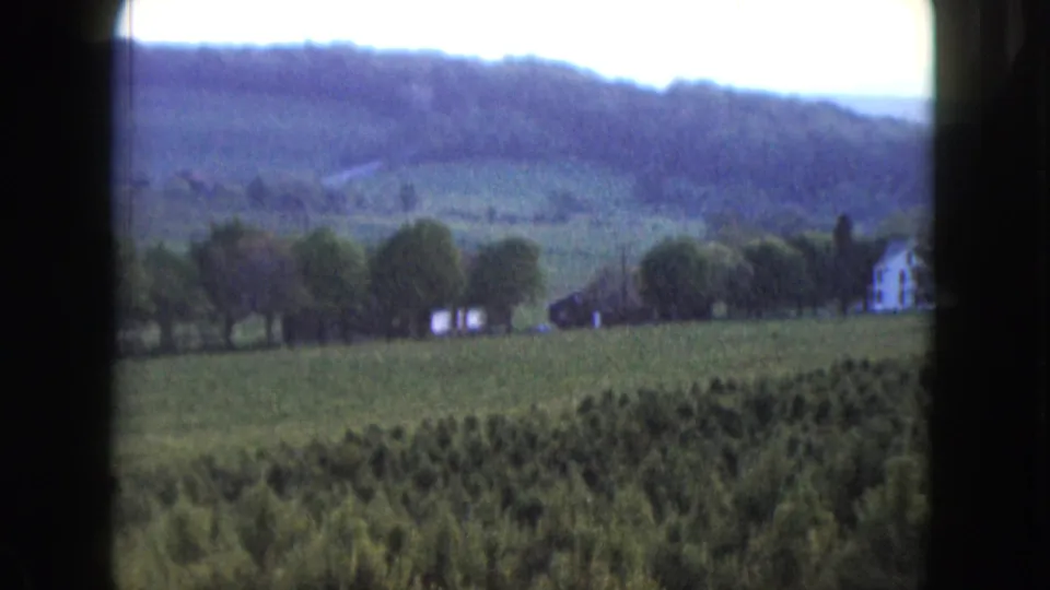1961 Nevada 8mm frame showing green rural fields, scattered trees, a white farmhouse, and wooded hills in the distance.