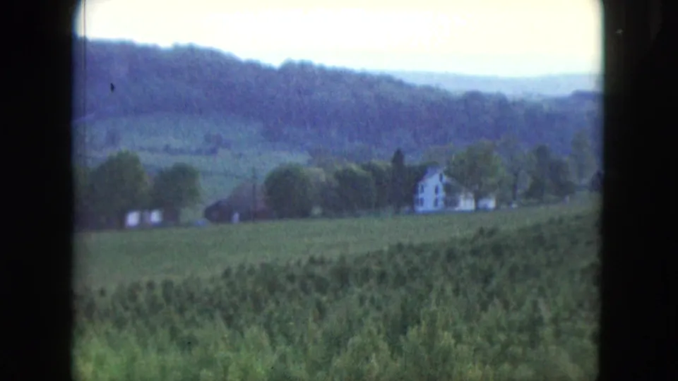 Soft 8mm frame from 1961 Nevada showing green fields, a farmhouse near trees, and hazy wooded hills.