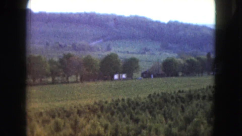 1961 Nevada 8mm frame of fields, trees, wooded hills, and small distant rural structures, with soft exposure and grain.