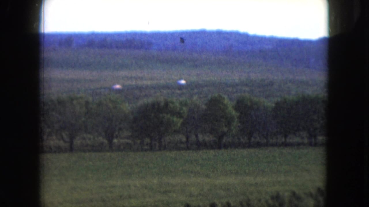 Rural field, tree line, and distant hills seen through dark film gate edges.