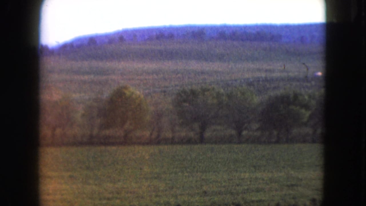 Rural landscape with field, trees, distant hills, pale sky, and dark vertical film edge.