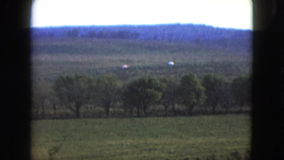 1961 Nevada 8mm frame showing a rural field and tree line with small cars crossing a distant road under low hills.