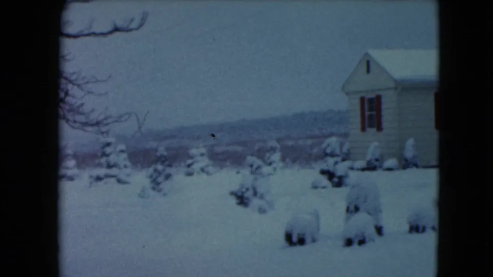 1961 8mm home-movie frame from Jamestown, Wisconsin showing a snowy yard, small evergreens, and a white house with red shutters.