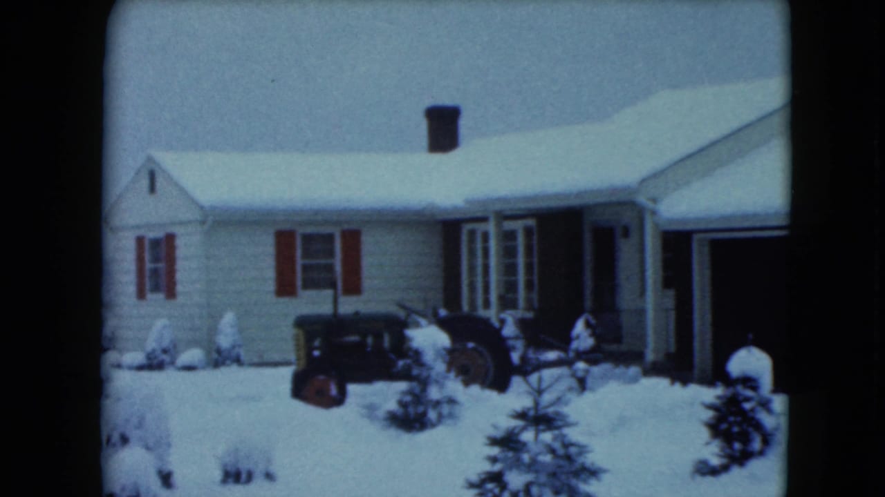 1961 Jamestown, Wisconsin home-movie frame showing a snow-covered house, red shutters, small evergreens, and a dark tractor in the yard.