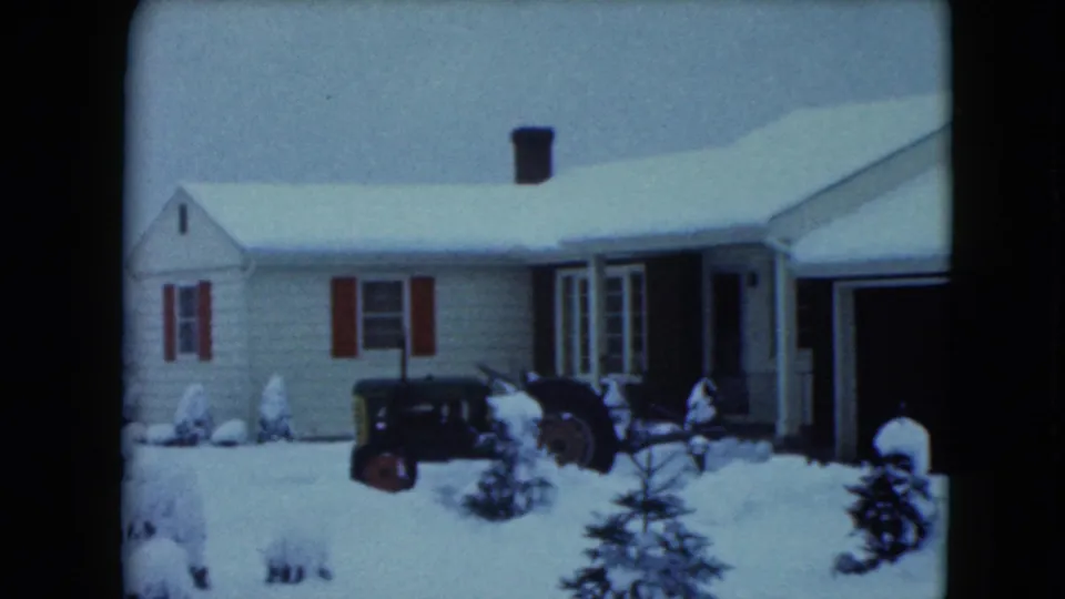 1961 Jamestown, Wisconsin home-movie frame showing a snow-covered house, red shutters, small evergreens, and a dark tractor in the yard.