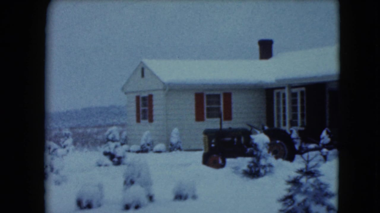 Snowy house exterior with red shutters, evergreens, and a tractor partly centered in the yard.