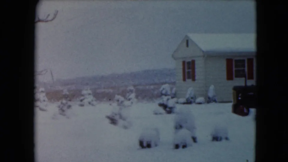 1961 Wisconsin frame near the end of the pan, with the house partly cropped, the tractor at right, and snow-covered shrubs before distant fields.
