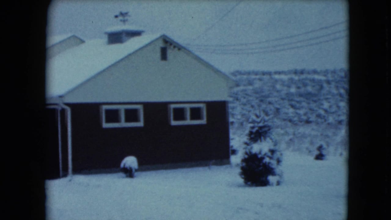 Snow-covered farmhouse side with garage wall, small windows, shrubs, and utility lines.