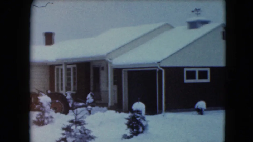 1961 Jamestown, Wisconsin 8mm view of the farmhouse front, attached garage, snowy yard, shrubs, and a tractor at the left edge.